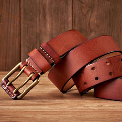 A brown cowhide leather belt with a copper buckle is coiled on a wooden surface, set against a rustic background. The durable belt features visible stitching near the buckle and multiple adjustment holes.