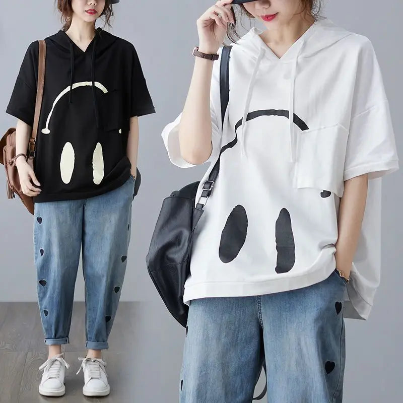 Two people wearing similar outfits: oversized Monday Mood hoodies with a large, abstract face print (one black, one white), blue jeans with small black heart patches, white sneakers, and carrying bags—perfect for adding spring colors to any look.