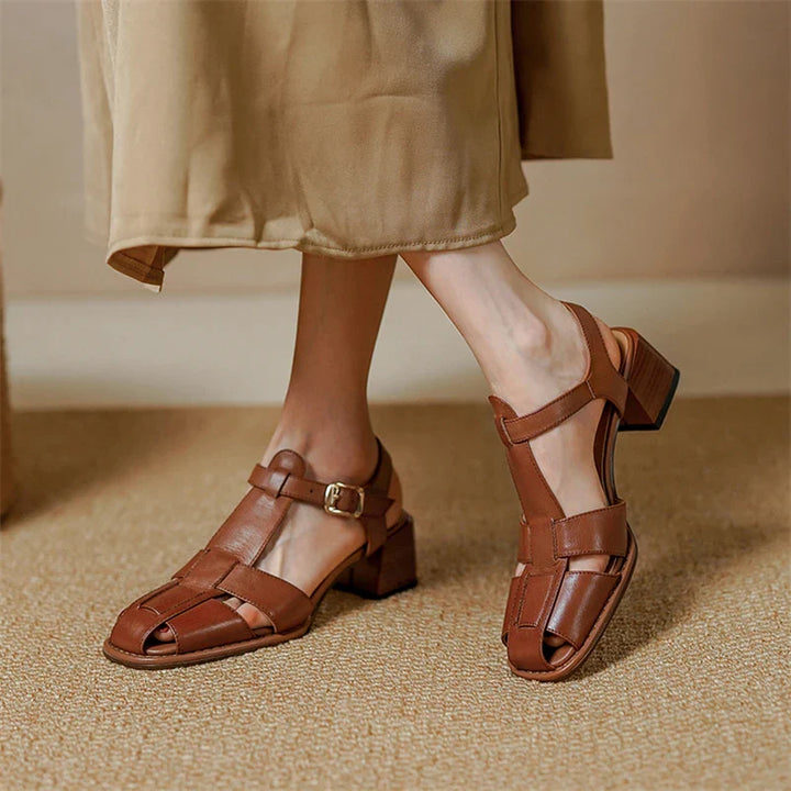 A person wearing a beige skirt and brown heeled sandals made from Genuine Italian Leather with ankle straps and an open toe design stands on a carpeted floor.
