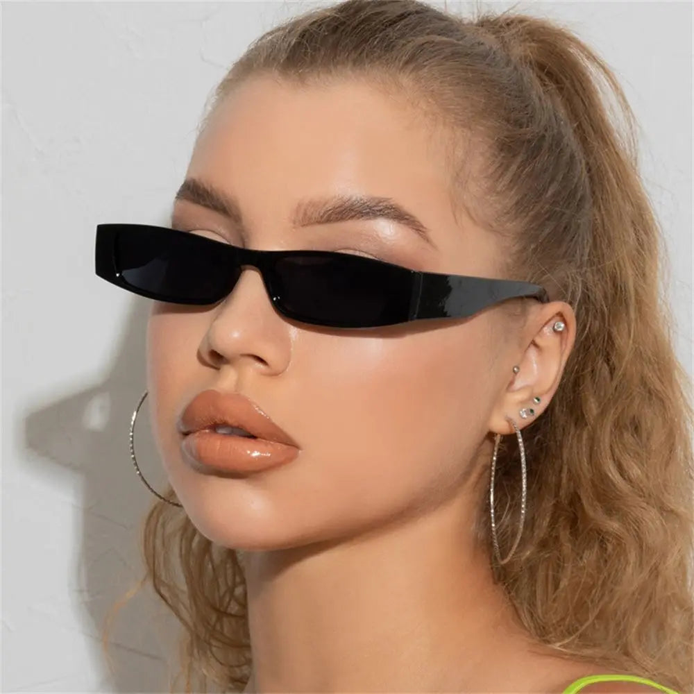 A woman with light skin and wavy blonde hair wears retro rectangle sunglasses with a lightweight frame, large hoop earrings, and glossy nude lipstick. She faces forward against a plain light background.