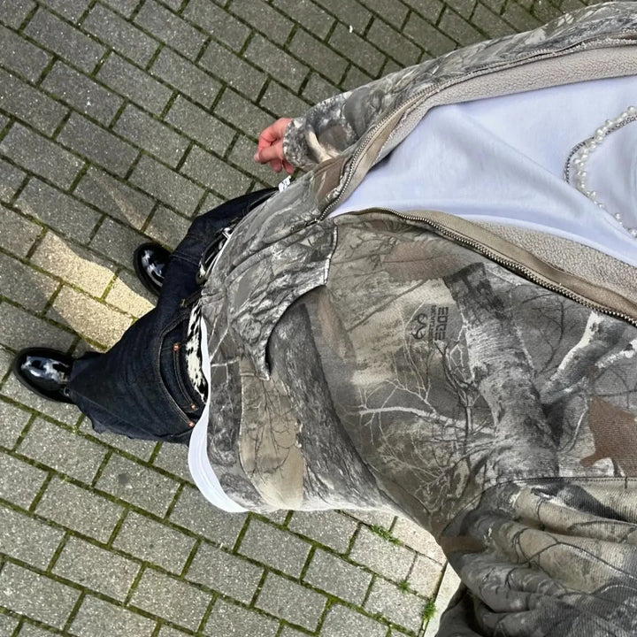A person wearing an oversized fit camouflage zip-up jacket, white shirt with a pearl necklace, dark pants, and black shoes stands on a paved brick sidewalk. The photo is taken from above, highlighting their stylish camo pattern outfit.