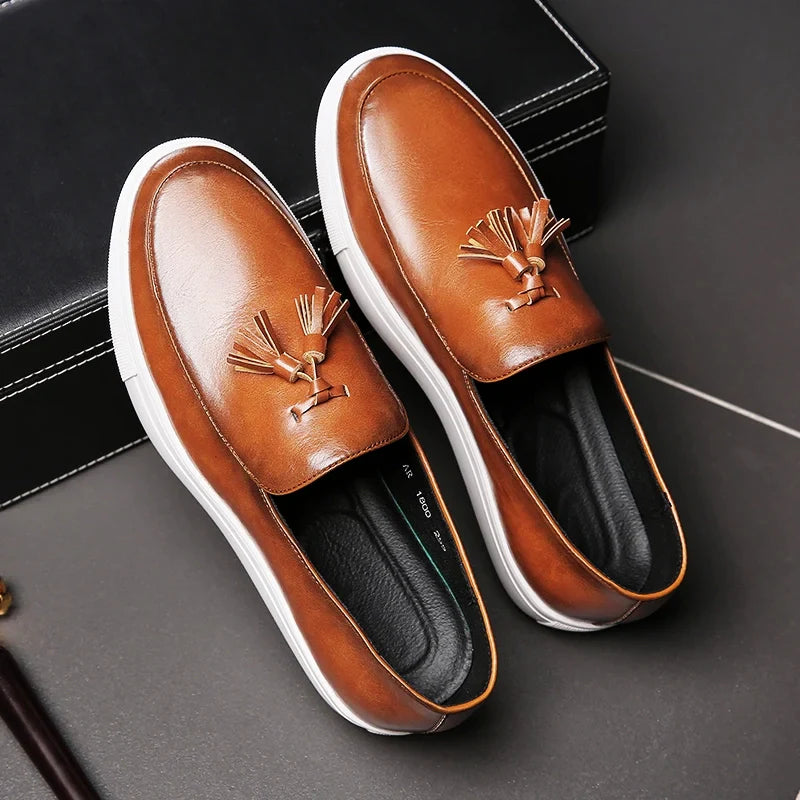 A pair of luxury leather loafers with white rubber soles and decorative tassels on the front, featuring a cushioned insole, placed on a dark surface next to a black box.