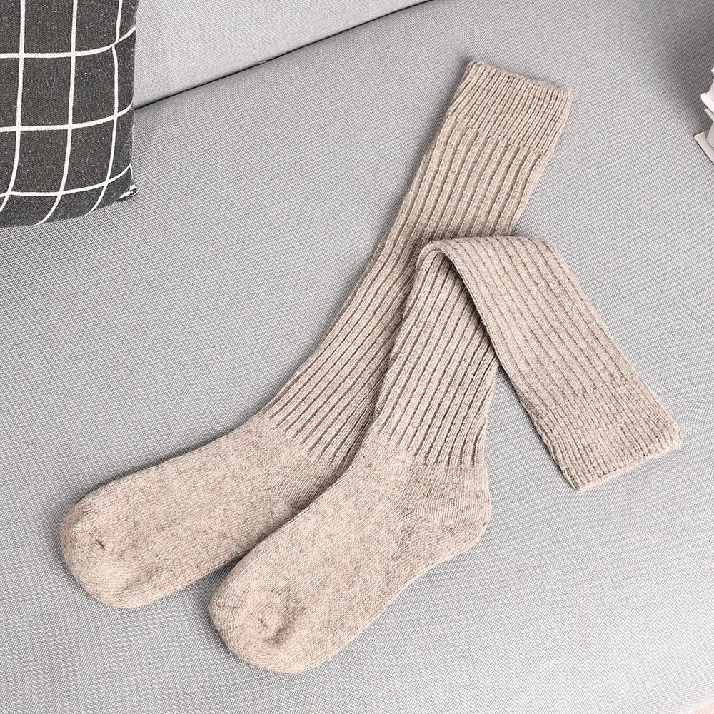 A pair of long, ribbed, beige knit socks made from a Cashmere cotton blend is laid out on a light gray couch next to a black and white checkered pillow.
