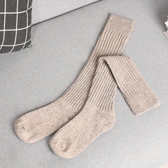 A pair of long, ribbed, beige knit socks made from a Cashmere cotton blend is laid out on a light gray couch next to a black and white checkered pillow.