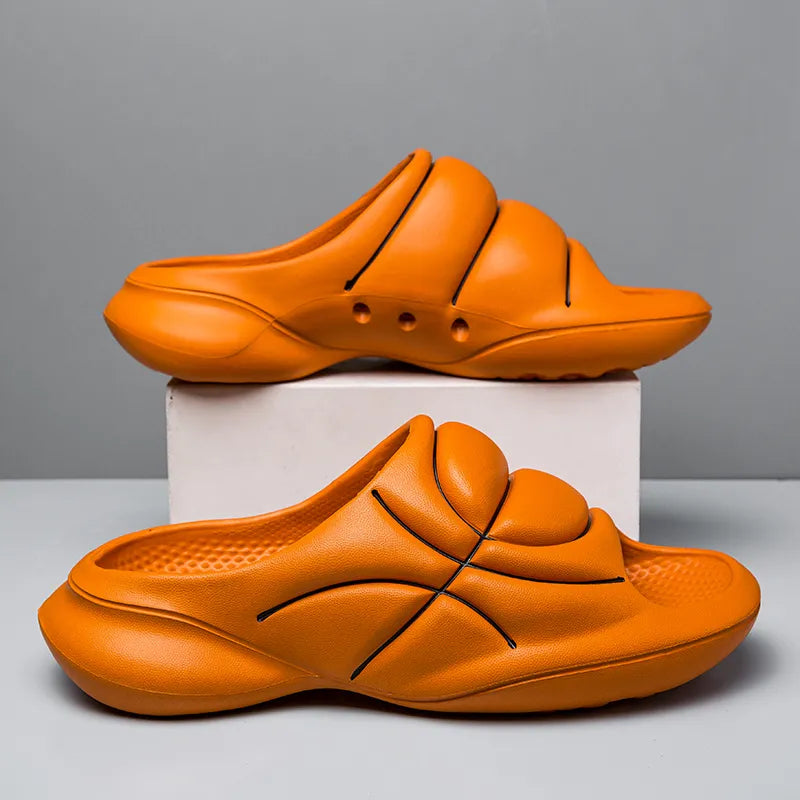Two orange basketball slip ons, designed with black lines like basketball seams, are displayed against a plain background. One sandal stands upright while the other rests on its side on a white block and both feature a cushioned footbed for comfort.