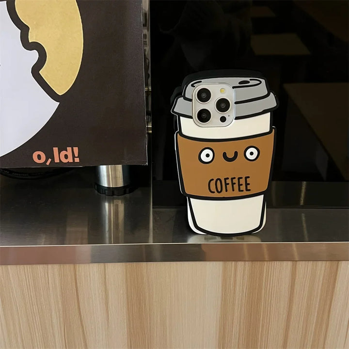 A phone with a 3D coffee cup soft silicone case sits on a counter next to a sign with part of a yellow and white design and the text o,Id! partially visible.
