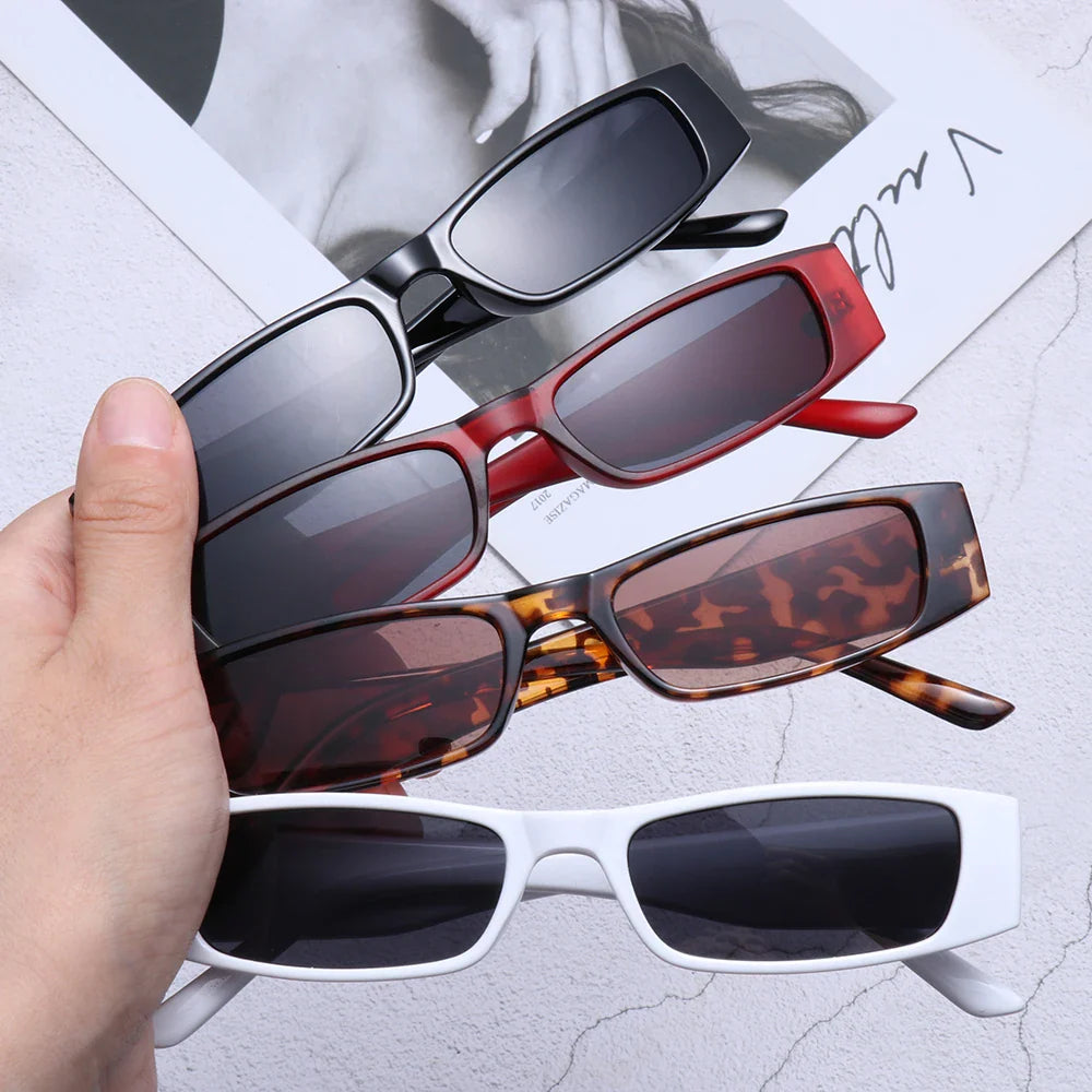 A hand holds four pairs of retro rectangle sunglasses—black, red, tortoiseshell, and white—with lightweight frames and a grayscale magazine page in the background.