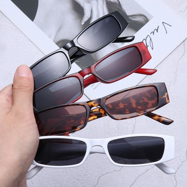 A hand holds four pairs of retro rectangle sunglasses—black, red, tortoiseshell, and white—with lightweight frames and a grayscale magazine page in the background.