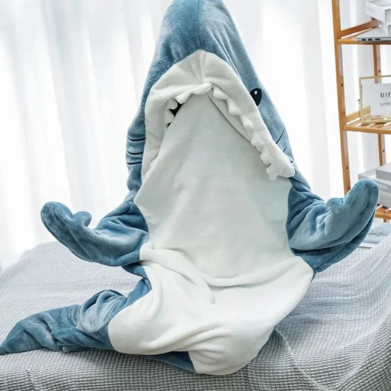 A person sits cross-legged on a bed wearing blue and white shark pajamas with the mouth open, their face peeking out playfully. A wooden shelf is in the background.