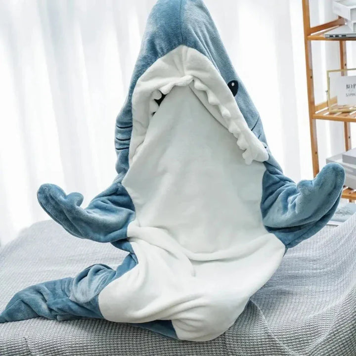 A person sits cross-legged on a bed wearing blue and white shark pajamas with the mouth open, their face peeking out playfully. A wooden shelf is in the background.