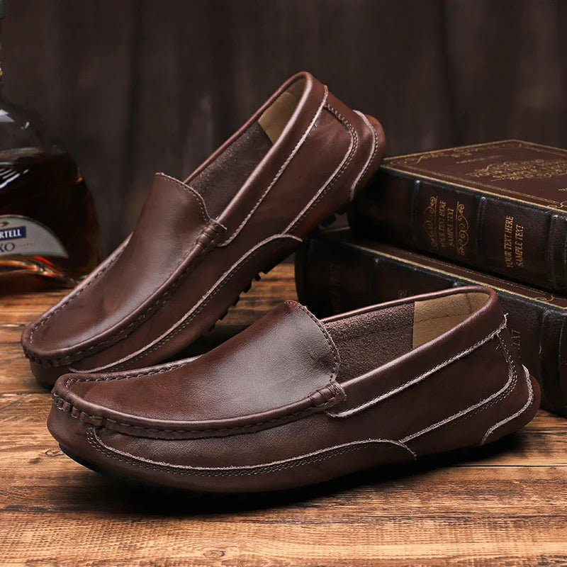 A pair of brown genuine leather loafers displayed on a wooden surface, with one shoe resting on books and the other lying flat. A glass bottle is partially visible in the background.