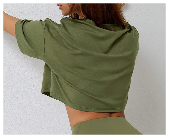 A woman with straight brown hair, wearing a relaxed fit cotton crop top and matching high-rise pants, stands with her back to the camera, one arm raised and touching a white wall.