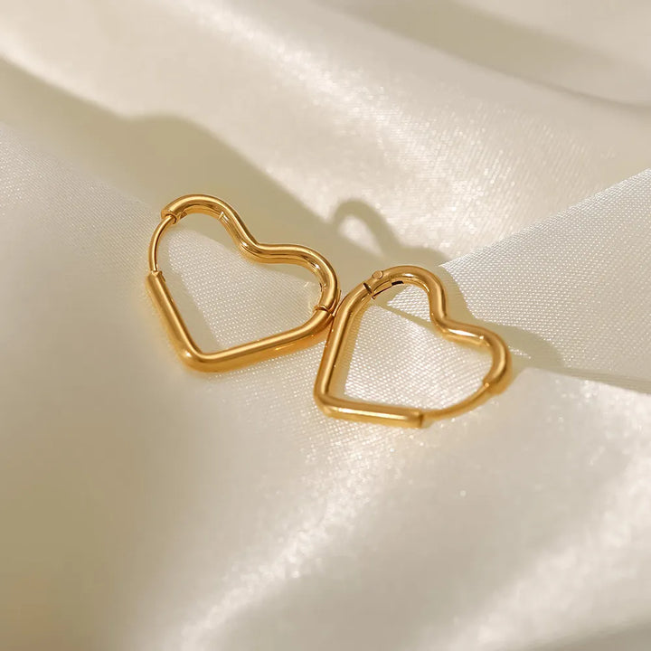 Two gold heart-shaped hoop earrings, known as the Eternal Love Earrings, rest on smooth, cream-colored satin fabric, catching the light and casting soft shadows—a perfect example of elegant craftsmanship.