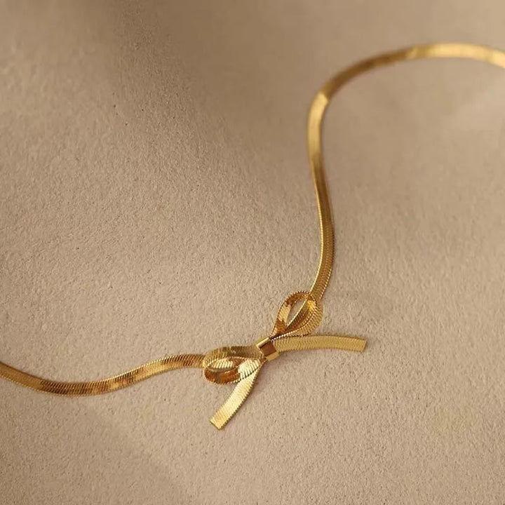 A thin, gold metallic ribbon tied in a small bow rests on a textured beige surface, evoking the refined simplicity of a minimalist jewelry bow necklace.