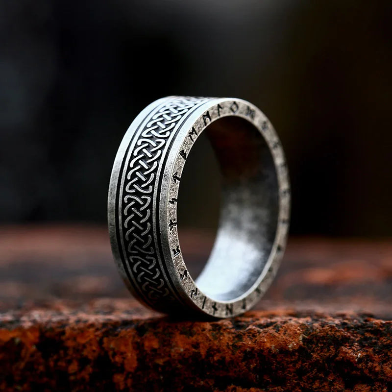 A durable metal Celtic knot ring with intricate patterns and ancient rune inscriptions sits upright on a rough, reddish-brown stone surface. The blurred background highlights the ring’s detailed engravings.