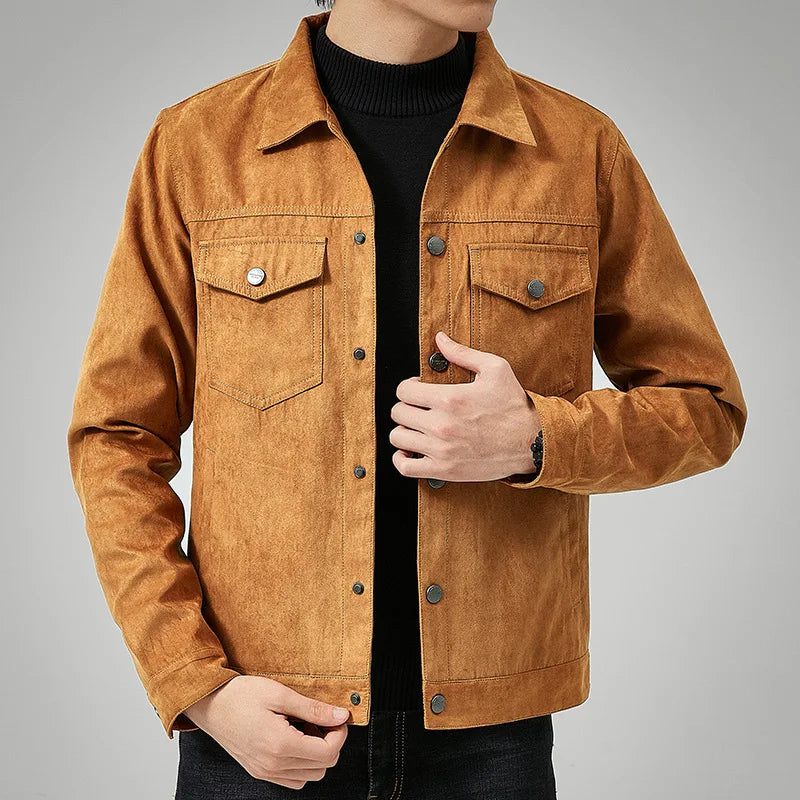 A person wearing a light brown suede jacket with a classic point collar over a black turtleneck, one hand adjusting the jacket and the other resting by their side, against a plain grey background.