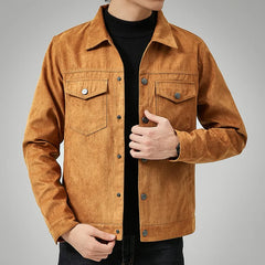 A person wearing a light brown suede jacket with a classic point collar over a black turtleneck, one hand adjusting the jacket and the other resting by their side, against a plain grey background.
