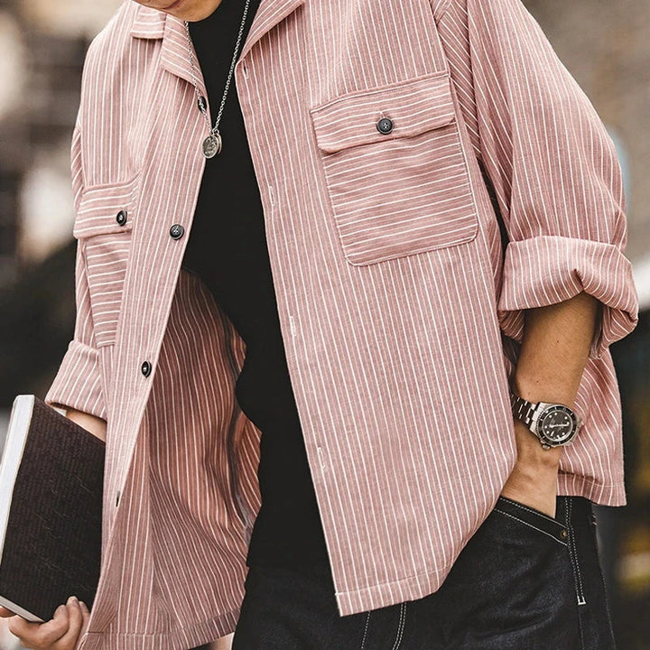 A person wearing a light pink and white pinstripe shirt with rolled-up sleeves, black shirt underneath, a watch on their wrist, and holding a closed notebook. Only the torso is visible.