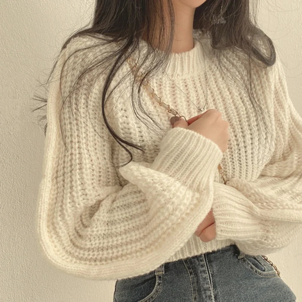A person wearing a chunky, cream-colored knit sweater with a ribbed texture and high-waisted blue jeans stands against a light background, their long hair down and one hand holding a chain strap over their shoulder.