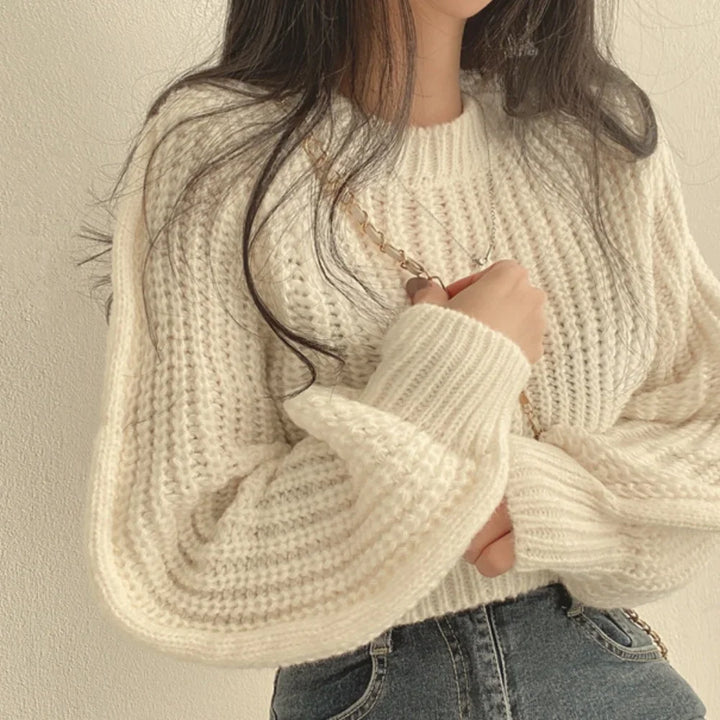 A person wearing a chunky, cream-colored knit sweater with a ribbed texture and high-waisted blue jeans stands against a light background, their long hair down and one hand holding a chain strap over their shoulder.