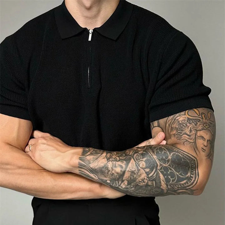 A man wearing a black quarter zip polo with ribbed detailing stands with his arms crossed, revealing a detailed tattoo sleeve on his right forearm, featuring various designs and a face.