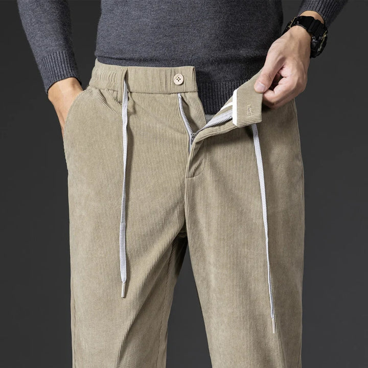 A person wearing men's smart casual corduroy pants with a drawstring, button, and zipper fly is unzipping the fly with one hand. They are also wearing a grey sweater and a black wristwatch.