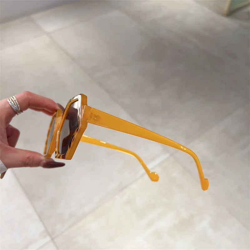 A hand holds yellow, hexagonal frame sunglasses with tinted lenses and UV400 protection, shown against a neutral tiled floor background.