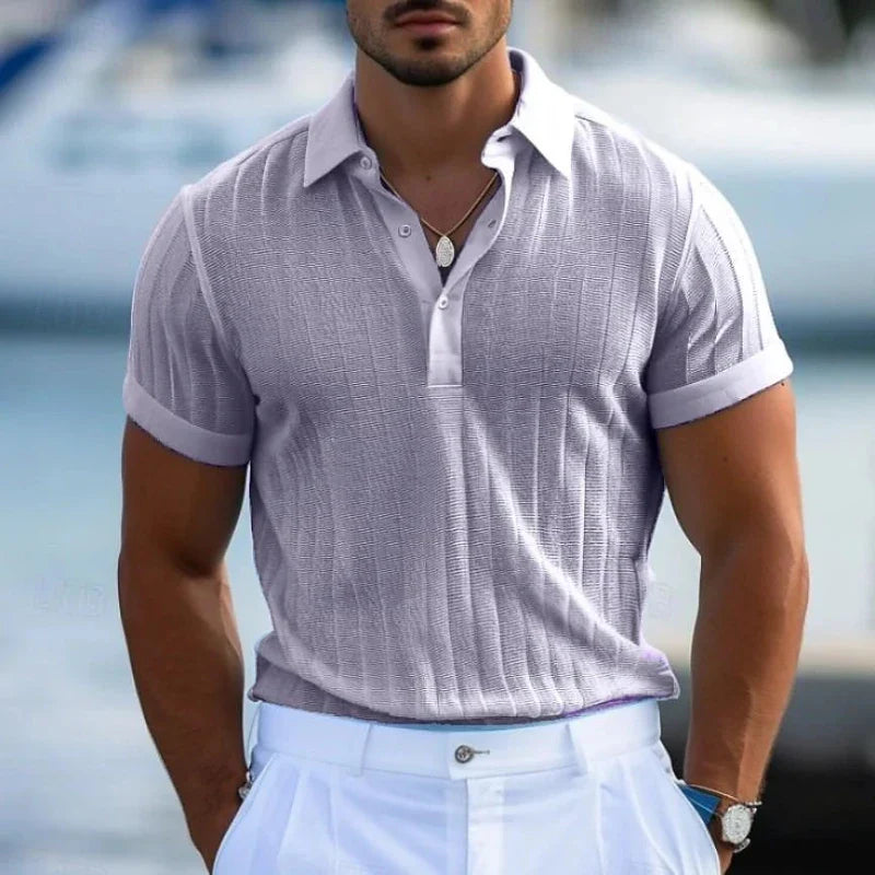 A man wearing a light purple short-sleeve polo in lightweight knit fabric and white pants stands outdoors. The background is blurred, suggesting a marina or waterfront setting. Only his upper body is visible.