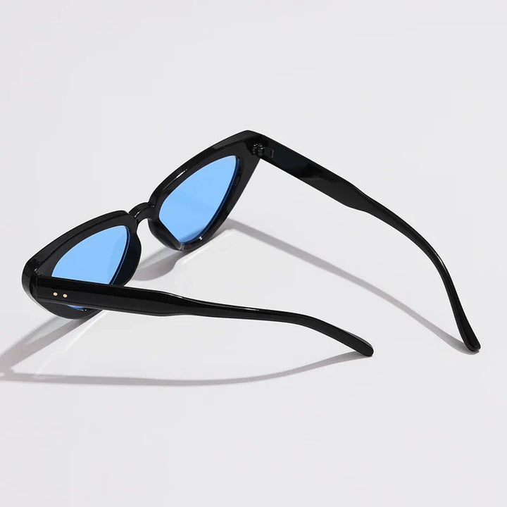 A pair of black-framed cat-eye sunglasses with blue-tinted lenses is placed on a white surface, the arms open and facing upward—perfect lightweight eyewear for style and comfort.