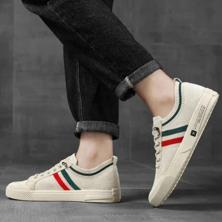 A person wearing beige casual sneakers with red and green stripes featuring a durable rubber outsole, paired with black jeans with rolled cuffs, stands on a grey floor against a textured grey background.