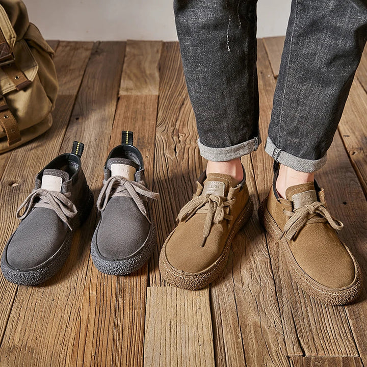 A person wearing rolled-up dark jeans stands on a wooden floor in tan cow suede leather Oxfords dress shoes. Next to them is an unworn pair of gray suede shoes, with a tan bag partially visible in the background.