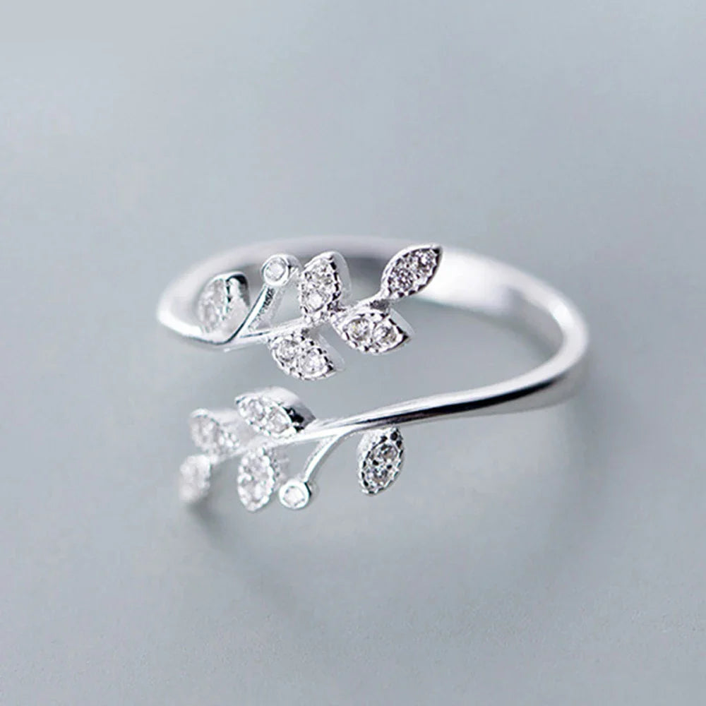 A silver Isadora Floral Ring featuring an open design with two branches adorned with small, sparkling leaf-shaped crystals on each end, set against a plain gray background—an elegant piece of fine jewelry.