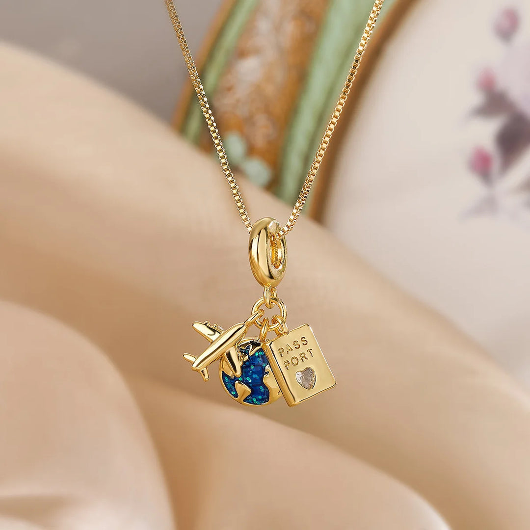 A gold adventure necklace features fine link craftsmanship with three charms—a small airplane, a blue-and-gold globe, and a rectangular passport charm with a tiny heart—set against a softly blurred beige and pastel background.