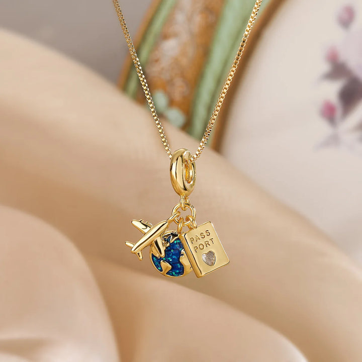 A gold adventure necklace features fine link craftsmanship with three charms—a small airplane, a blue-and-gold globe, and a rectangular passport charm with a tiny heart—set against a softly blurred beige and pastel background.