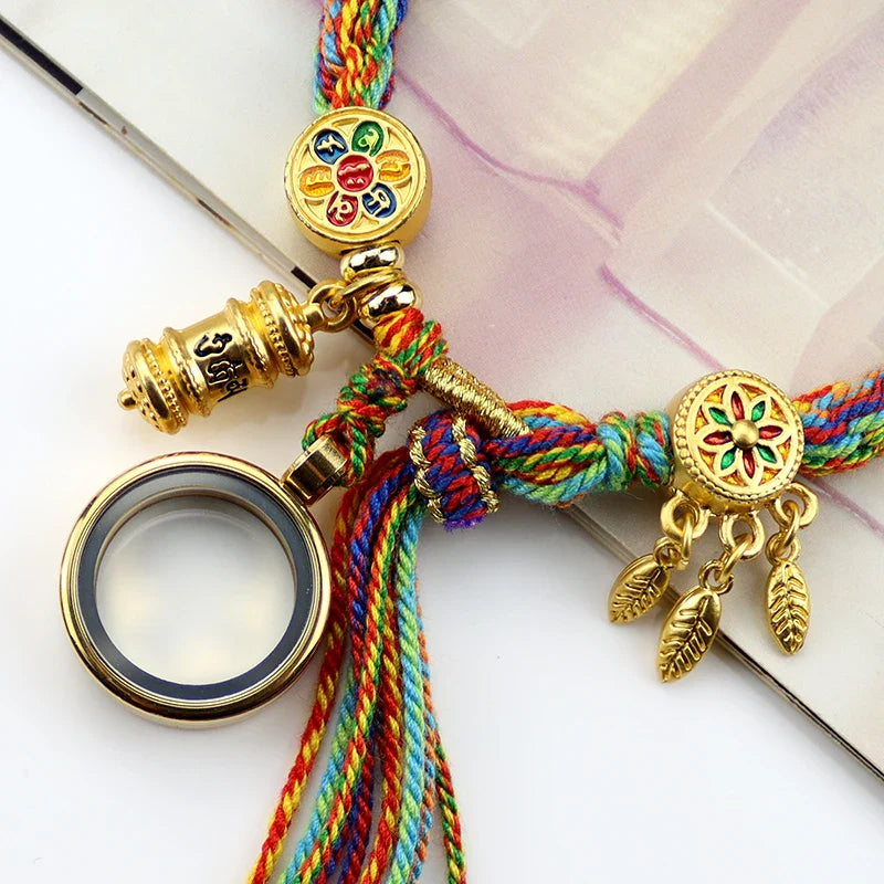 Colorful braided string bracelet featuring gold charms—a magnifying glass, a spinning prayer wheel inspired by Tibetan craftsmanship, and a floral charm with dangling gold feather pendants—resting on a book.