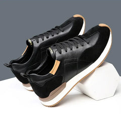 A pair of stylish black sneakers with white soles and brown accents are displayed on a white geometric platform against a gray background. Crafted with top-grain leather and suede, these shoes offer both elegance and arch support.