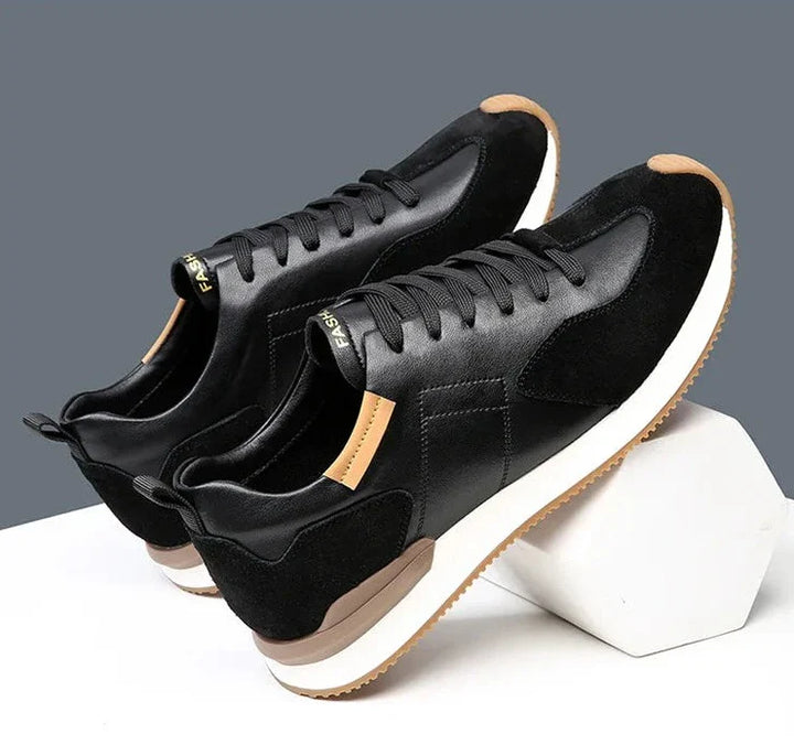 A pair of stylish black sneakers with white soles and brown accents are displayed on a white geometric platform against a gray background. Crafted with top-grain leather and suede, these shoes offer both elegance and arch support.