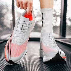 A person wearing a white sock holds up one white and orange running shoe featuring a shock-absorbing sole while the other shoe is worn, standing on a black textured surface with a blurred background.