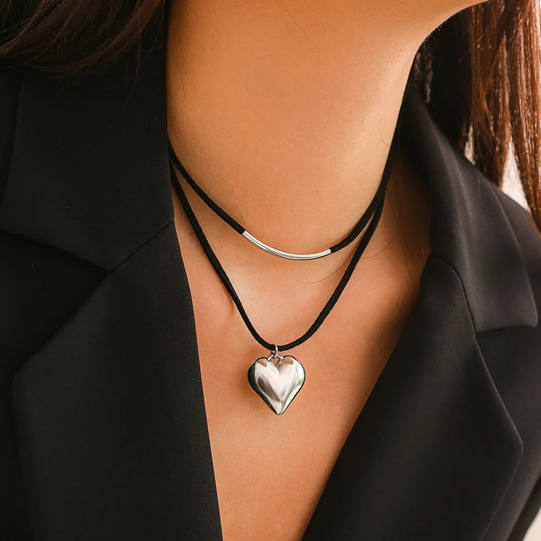 A person wearing a black blazer showcases two black cord necklaces: an adjustable choker with a silver bar and another featuring a shiny vintage heart pendant. The photo focuses on their neck and collar area.
