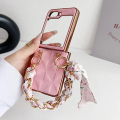 A hand holds a pink Samsung Galaxy Z Flip premium leather case with a quilted pattern, built-in mirror, shockproof protection, and a decorative chain handle wrapped with a floral scarf. A white wall and partially visible book are in the background.