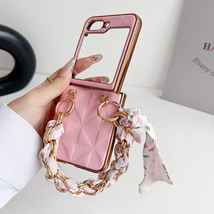 A hand holds a pink Samsung Galaxy Z Flip premium leather case with a quilted pattern, built-in mirror, shockproof protection, and a decorative chain handle wrapped with a floral scarf. A white wall and partially visible book are in the background.