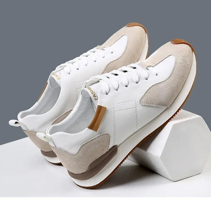 A pair of stylish white sneakers with top-grain leather, light beige suede accents, and brown rubber soles offer excellent arch support, displayed on white geometric blocks against a gray background.