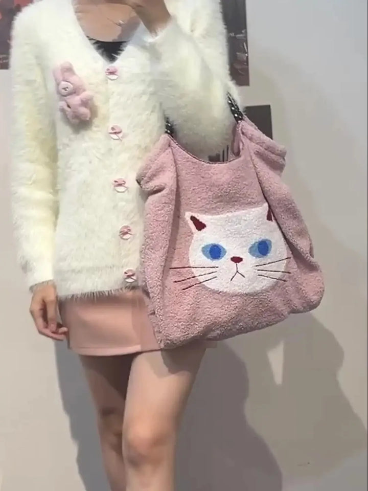 A person wearing a fuzzy white cardigan with pink buttons and a pale pink skirt holds an animal pattern tote—a large pink bag featuring a white cat face with blue eyes.