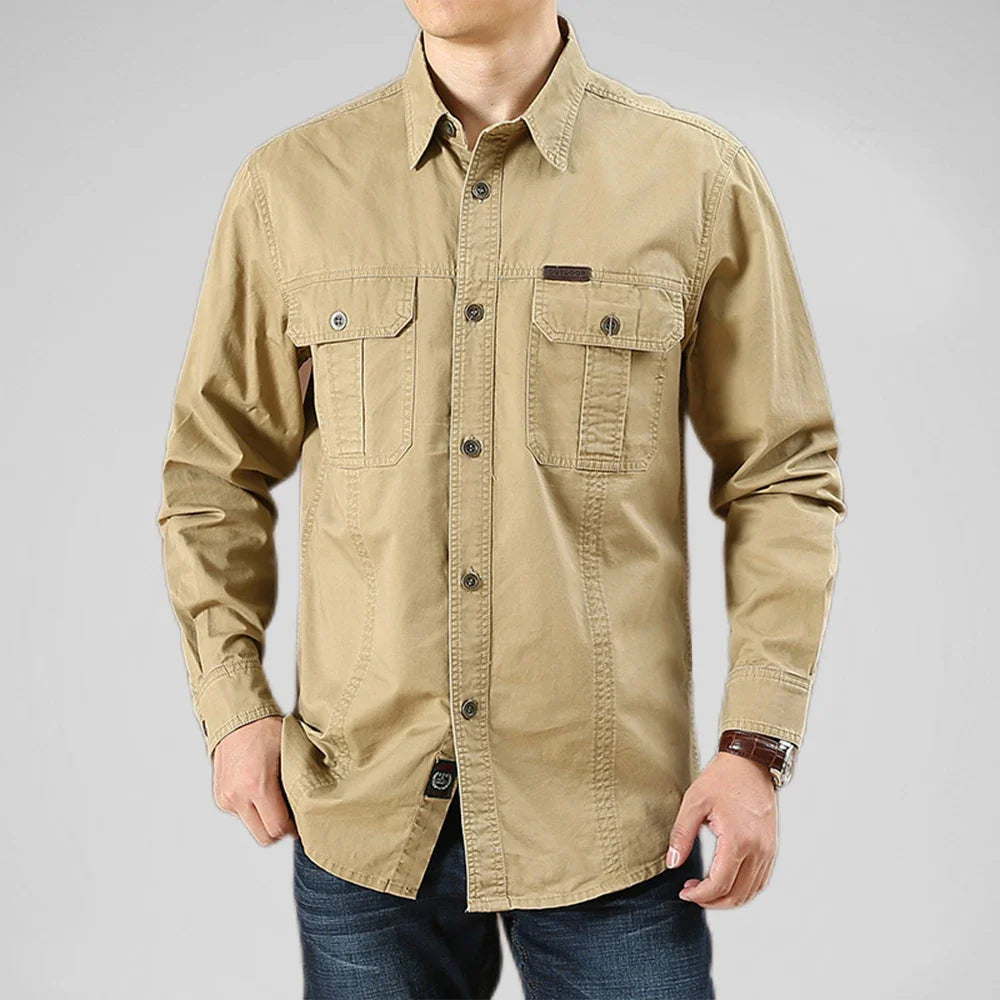 A man wearing a light brown 100% cotton shirt with two chest pockets and dark jeans, standing against a plain light gray background. His face is not visible.