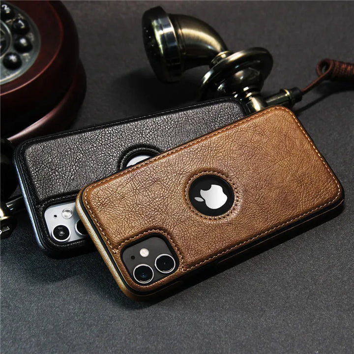 Two iPhones with luxury leather phone cases—one brown, one black—rest on a dark surface beside an old-fashioned rotary handset. Each slim protective case features a circular cutout for the Apple logo and is wireless charging compatible.