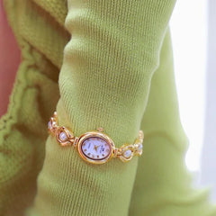 A gold bracelet watch with precision quartz movement and pearl accents is worn over a light green, ruffled long-sleeve top. The small, oval watch face displays Roman numerals.