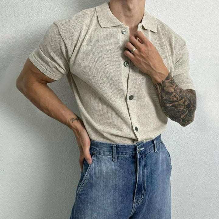 A person wears a short-sleeve unisex top and blue jeans, standing against a light wall. One arm rests on their hip while the other hand touches their collar, revealing a tattoo sleeve on their forearm.