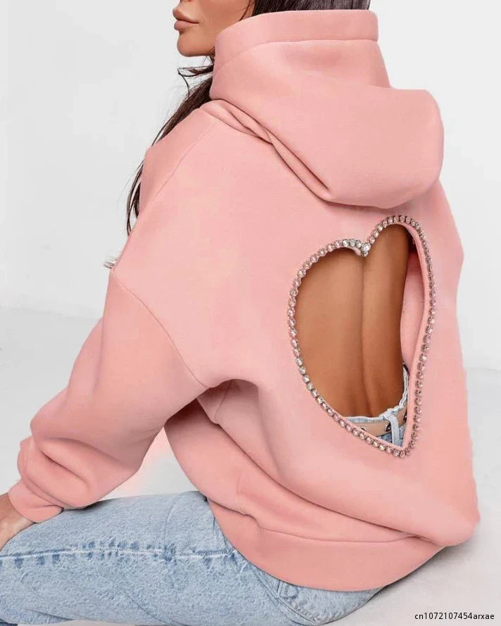 A woman wears a light pink heart-shaped open back hoodie with a rhinestone trim, revealing her bare skin. She pairs the oversized hoodie with light blue jeans and is seated, facing away from the camera.