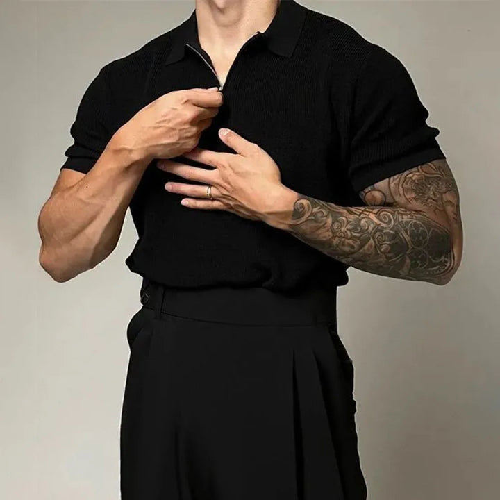 A man wearing a short-sleeved black quarter zip polo and high-waisted black trousers adjusts his collar. His left arm, covered in a tattoo sleeve, crosses his chest as he uses his right hand to zip up the shirt.