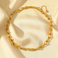 A gold statement chain necklace with elongated links and small round beads, crafted from durable 316L stainless steel and displayed on a soft cream-colored fabric background.